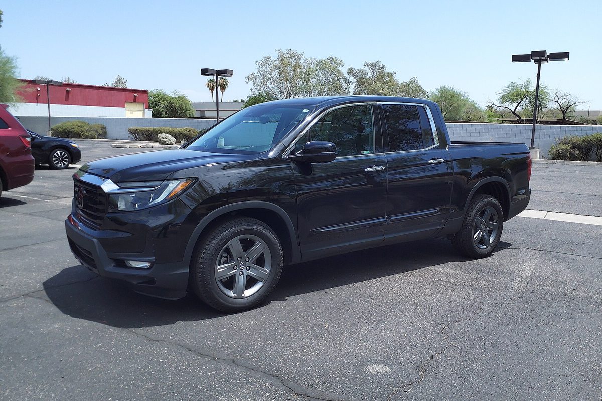 A black 2023 Honda Ridgeline RTL-E pickup parked in a sunny lot, with a red building and trees behind it