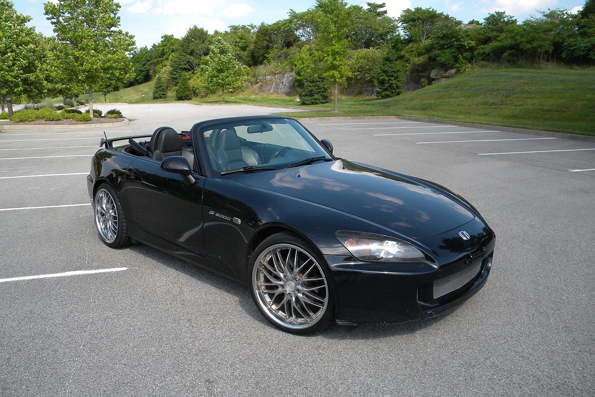 A black 2007 Honda S2000 with its top down and multi-spoke chrome wheels, parked alone in an empty lot below a grassy, tree-lined hill
