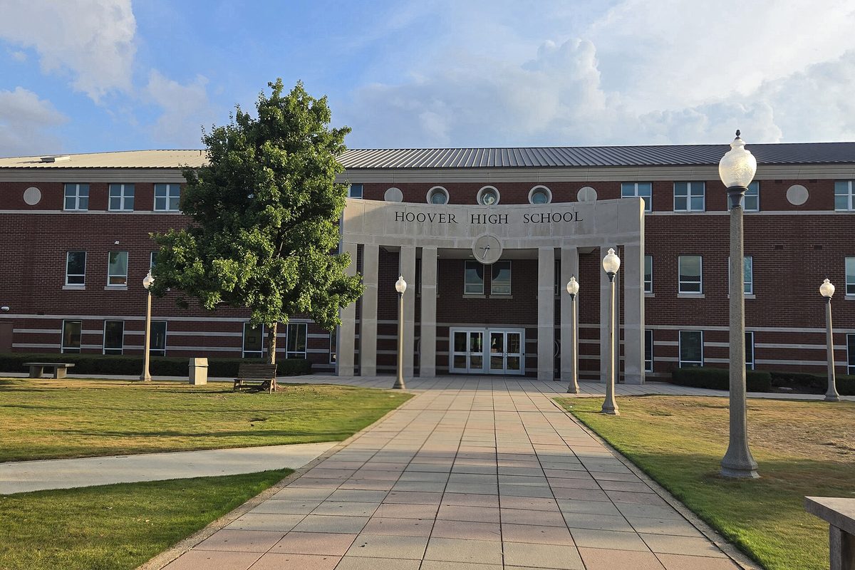 The red brick front of Hoover High School with its columned entrance, a paved walkway lined with lamp posts and a leafy tree on the lawn