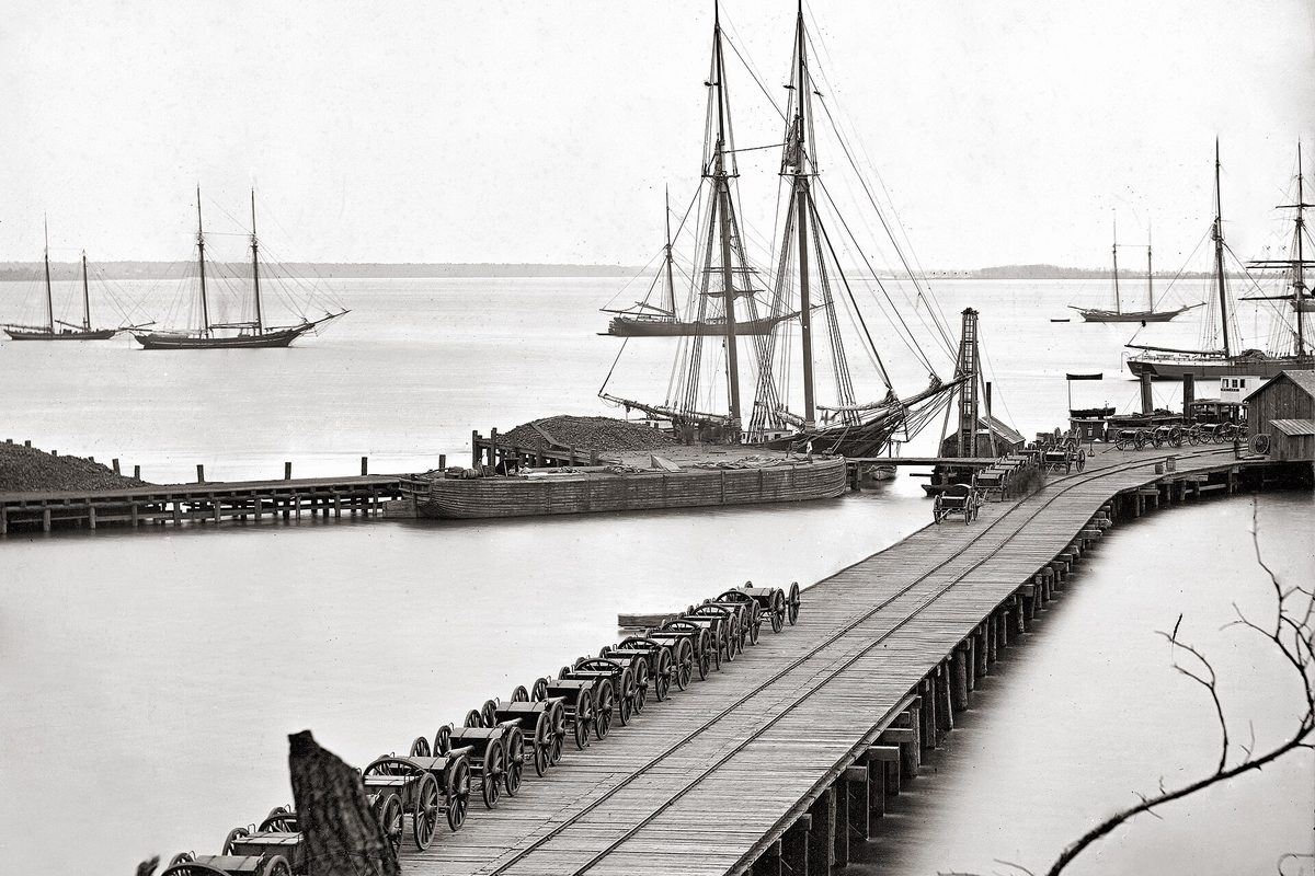 Civil War photograph of the City Point waterfront: a long wooden wharf with a rail track, a row of wheeled gun carriages, and sailing schooners on the river
