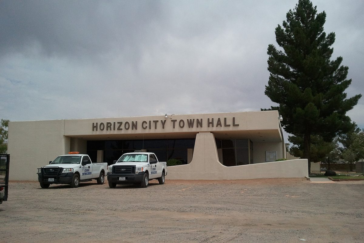 Horizon City Town Hall, a low tan stucco building with its name in large letters, two white city pickup trucks parked on the gravel lot and a tall pine under a stormy sky