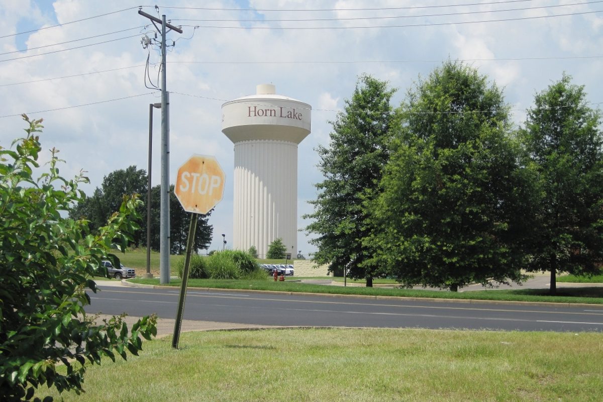The white Horn Lake water tower behind a row of trees, with a stop sign and a four-lane road in front