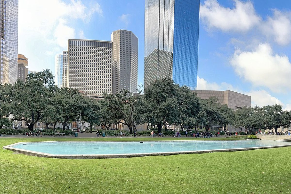 The long reflecting pool and live oaks of Hermann Square in downtown Houston, with glass and stone office towers rising behind them on a sunny day