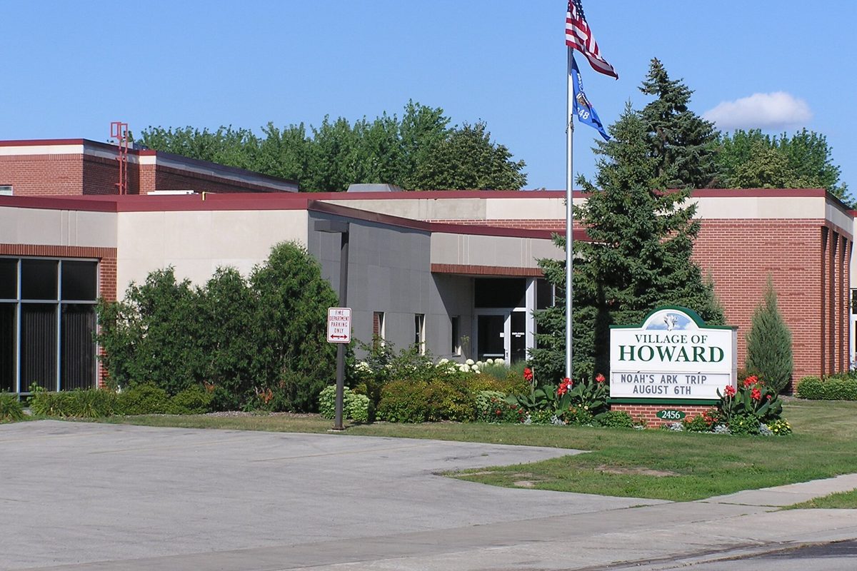 Howard Village Hall, a low brick and stucco building with a Village of Howard sign, flower beds and flags out front
