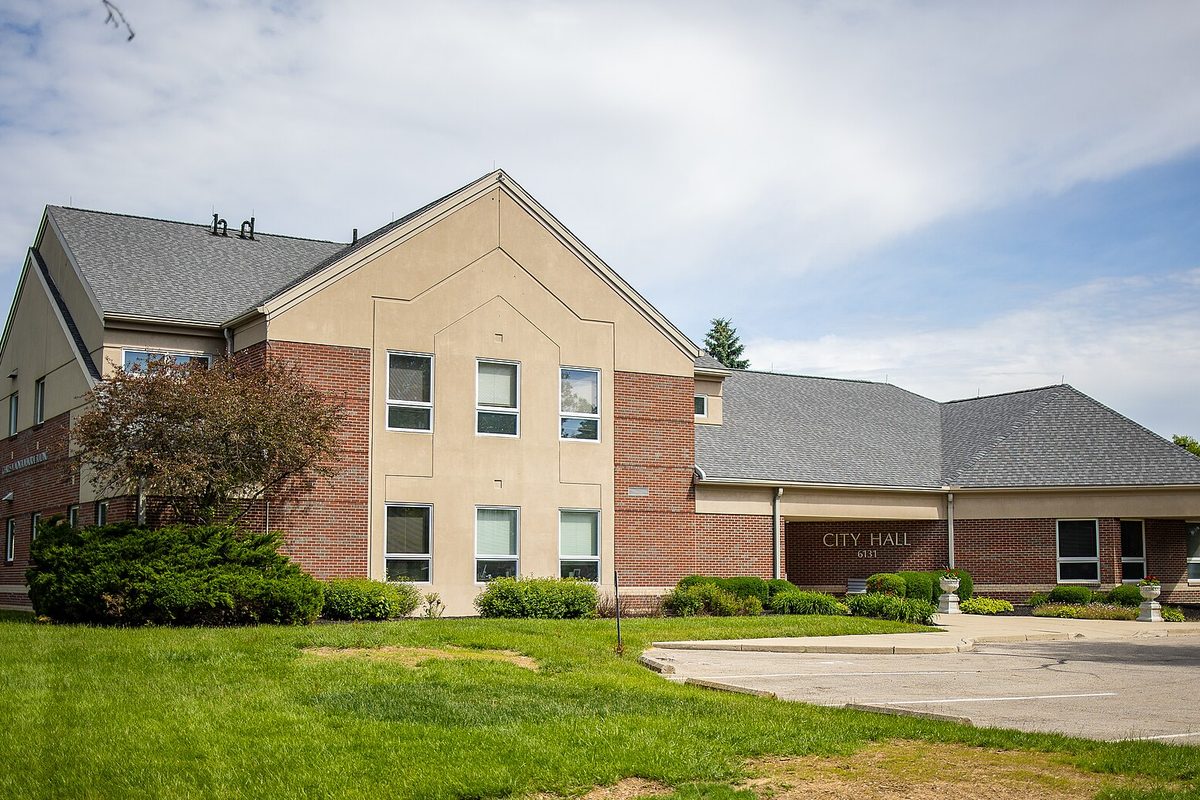Huber Heights City Hall, a two-story brick and tan building with a gabled front, a lettered City Hall sign, a lawn and a curving drive