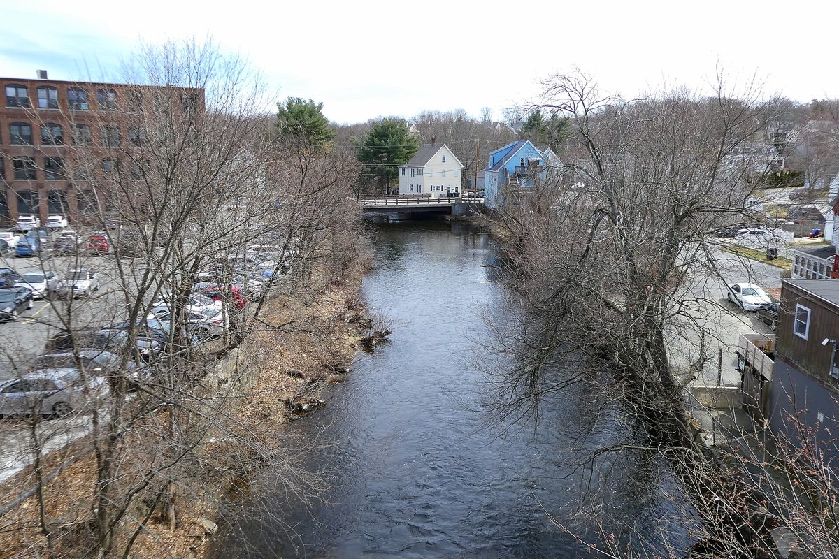 The Assabet River running through Hudson between bare trees, a parking lot and brick mill building on the left and a bridge with houses upstream