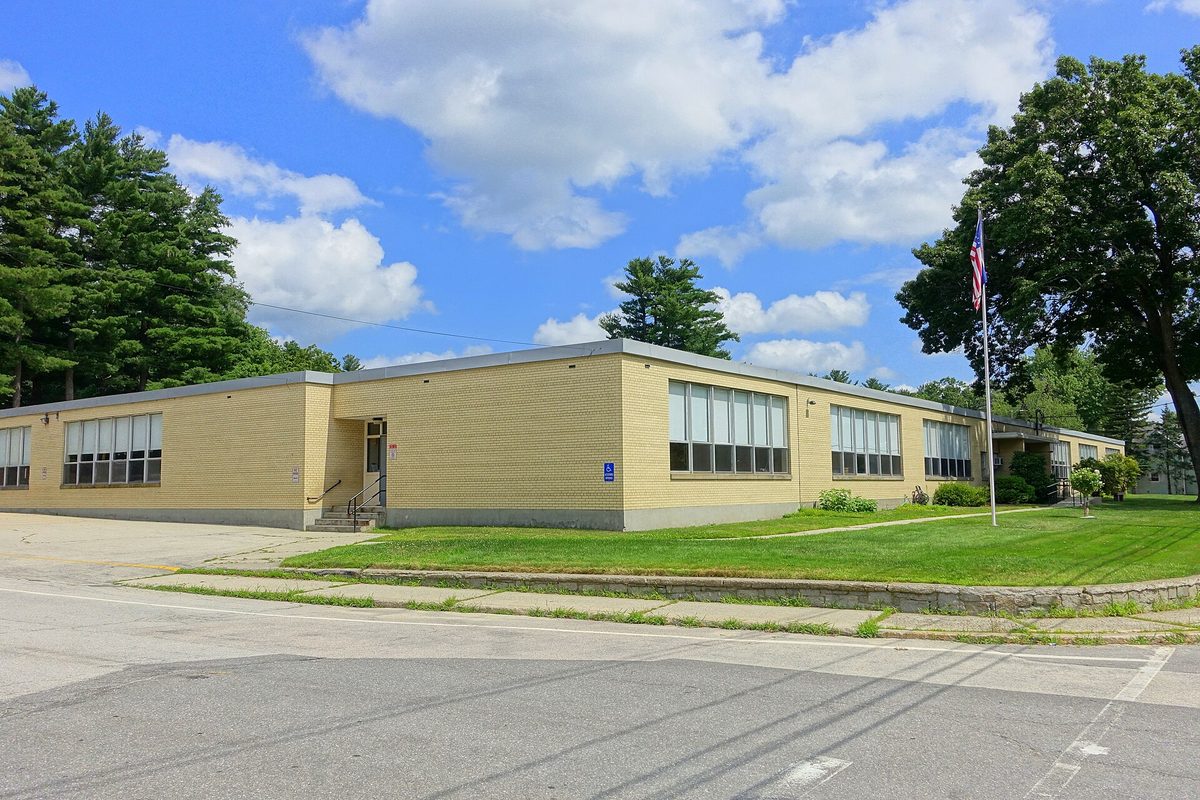 The low yellow-brick Library Street School in Hudson, with a flagpole, a green lawn and tall pines under a cloudy blue sky