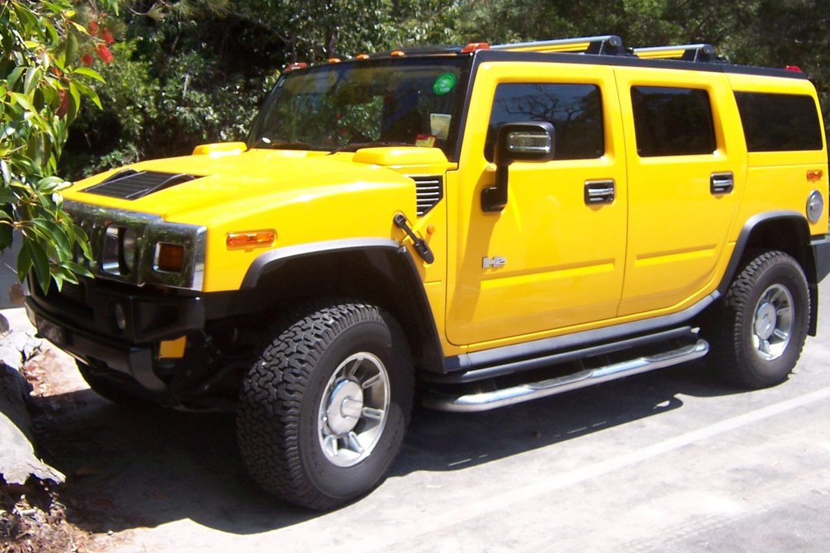 A bright yellow Hummer H2 SUV with chrome wheels, side steps and a roof rack, parked on a paved road under trees