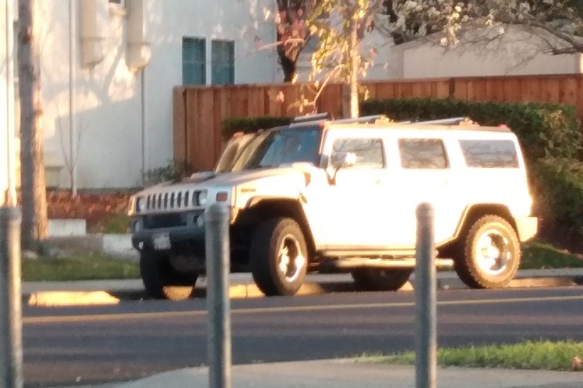 A white Hummer H2 parked on a street in front of a wooden fence and a pale building, lit by low evening sun