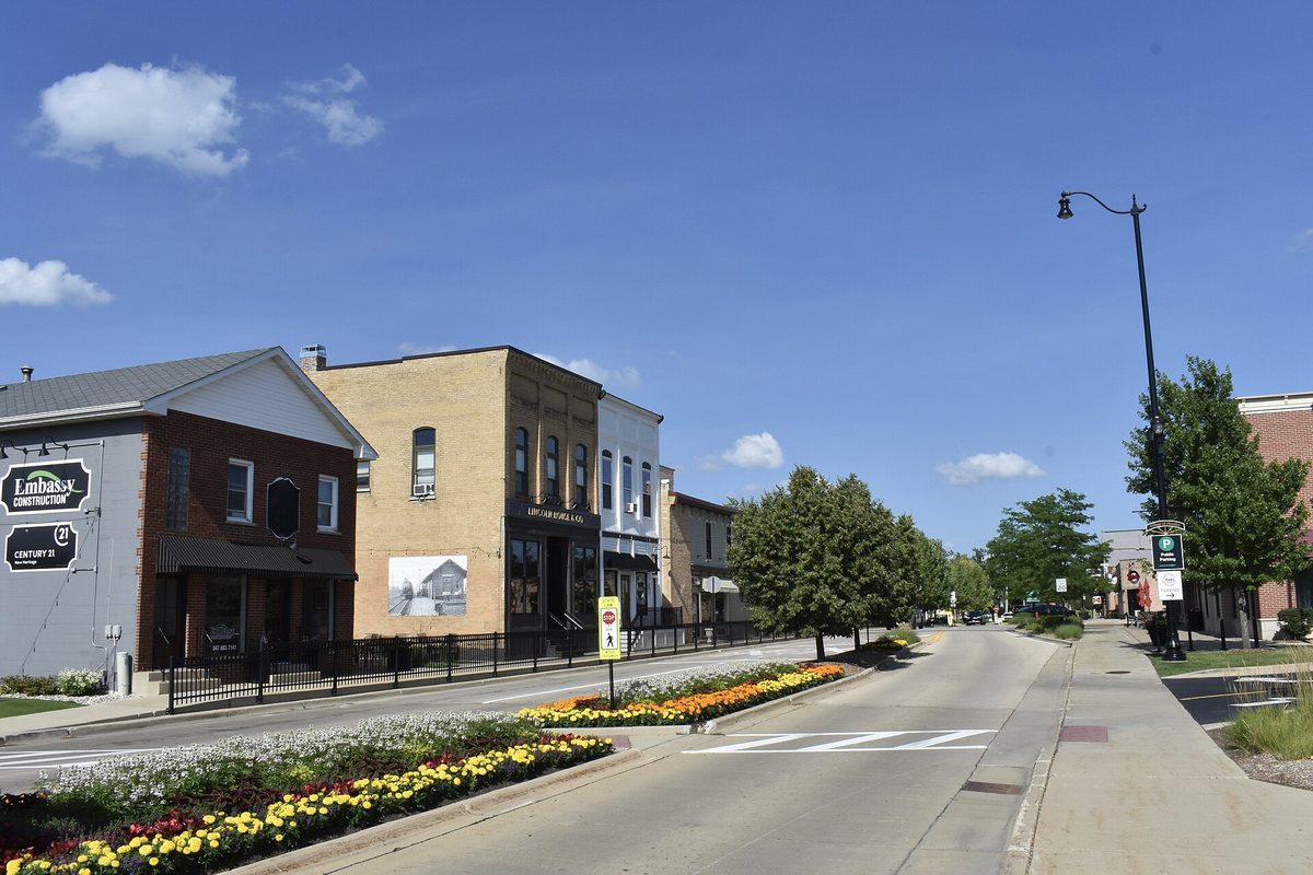 Downtown Huntley on a sunny day, with brick and stone storefronts, a lamppost and beds of yellow and red flowers along the street