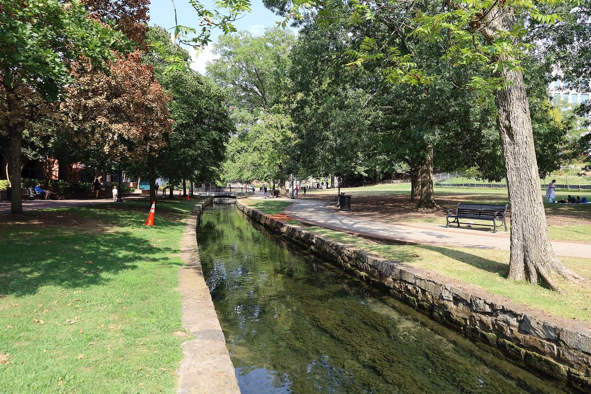 A clear, stone-lined stream channel running under tall shade trees in Big Spring International Park, with a bench and a paved path beside it