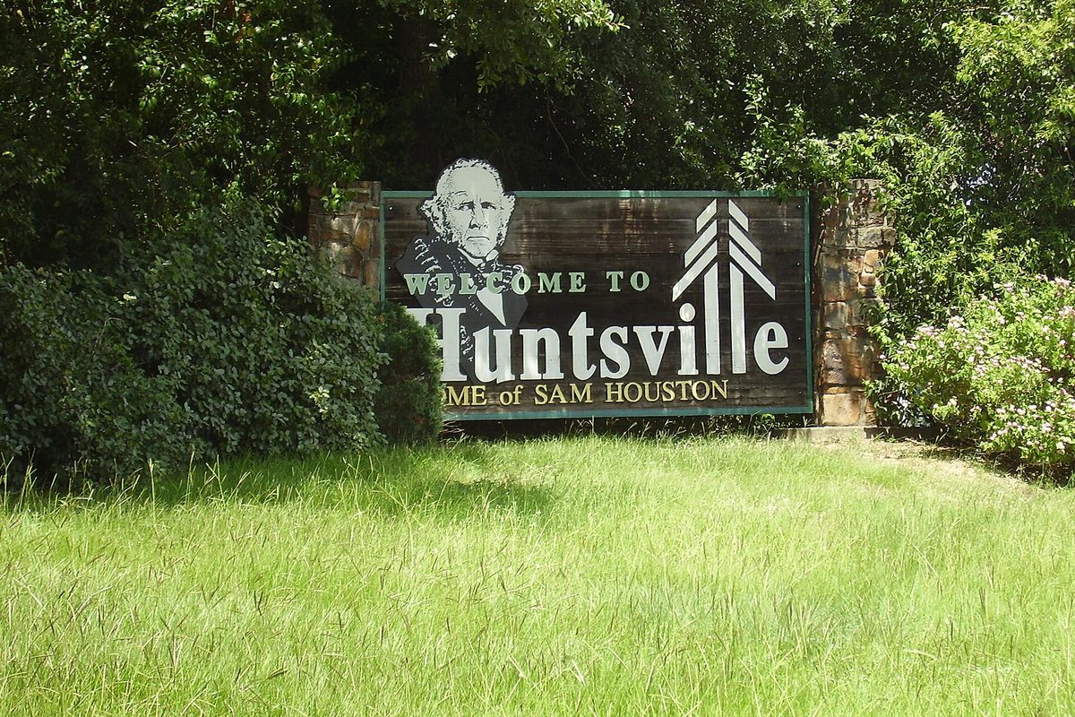 A wooden Welcome to Huntsville, Home of Sam Houston sign with Sam Houston's portrait, set in stone pillars among shrubs above tall green grass