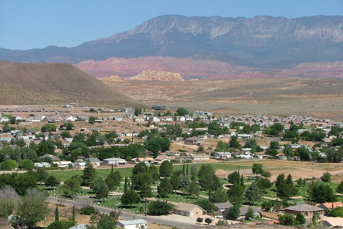 Homes and green trees in Hurricane, Utah, below a brown hill, pink red-rock cliffs and a long gray mesa under a clear sky