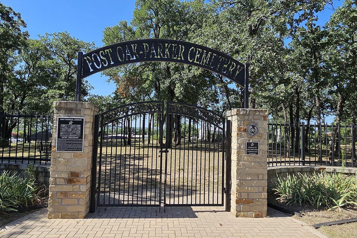 The arched iron gate of Post Oak-Parker Cemetery in Hurst, lettered with its name and set between tan stone pillars, with oak trees behind