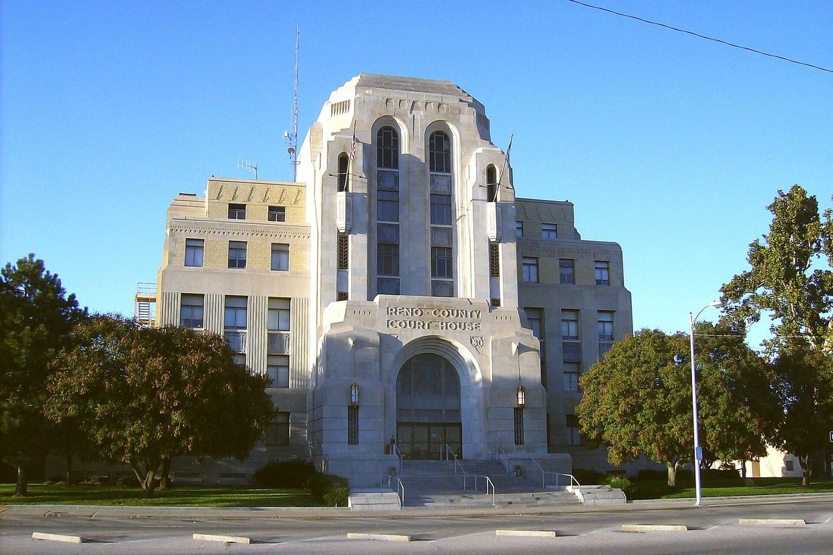 The pale stone Reno County Courthouse in Hutchinson, a tall art deco tower over a carved arched entrance, framed by trees under a clear blue sky