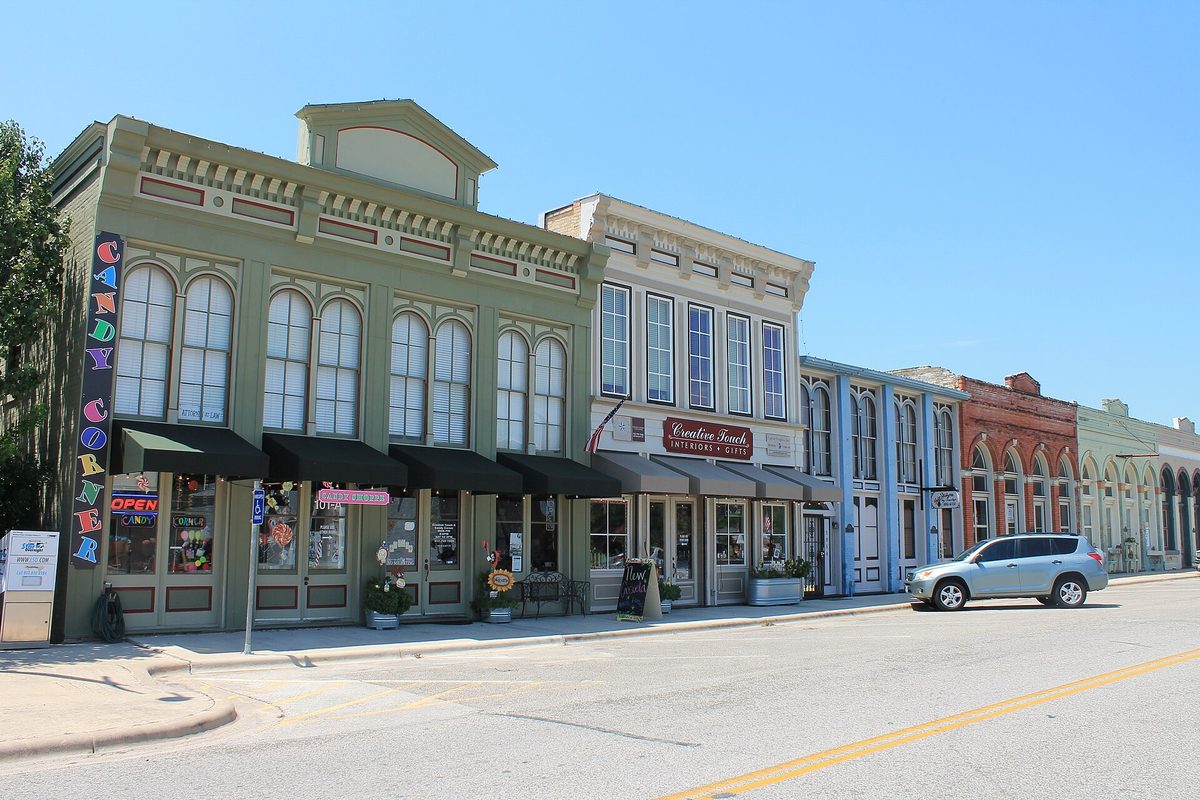 A row of old two-story storefronts in Hutto's historic commercial district, led by a green building with tall arched windows and a colorful vertical candy shop sign, with a silver SUV parked on the street