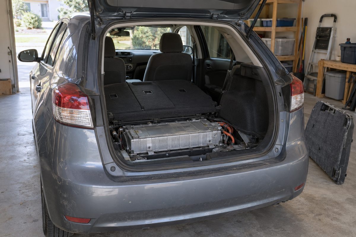 A gray hatchback in a garage with its rear hatch open and the cargo floor lifted out, showing the hybrid battery pack and its orange cables