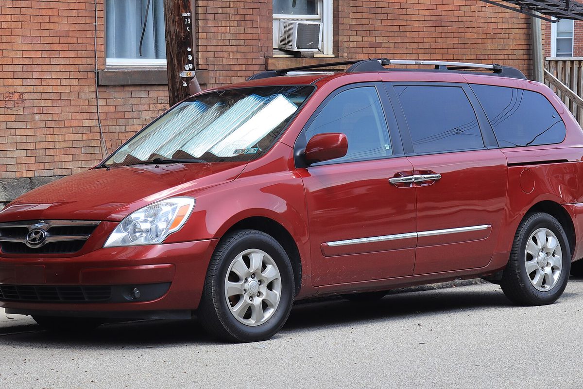 A cranberry red 2008 Hyundai Entourage minivan, front left view, parked on a street by a brick building with a sunshade in the windshield