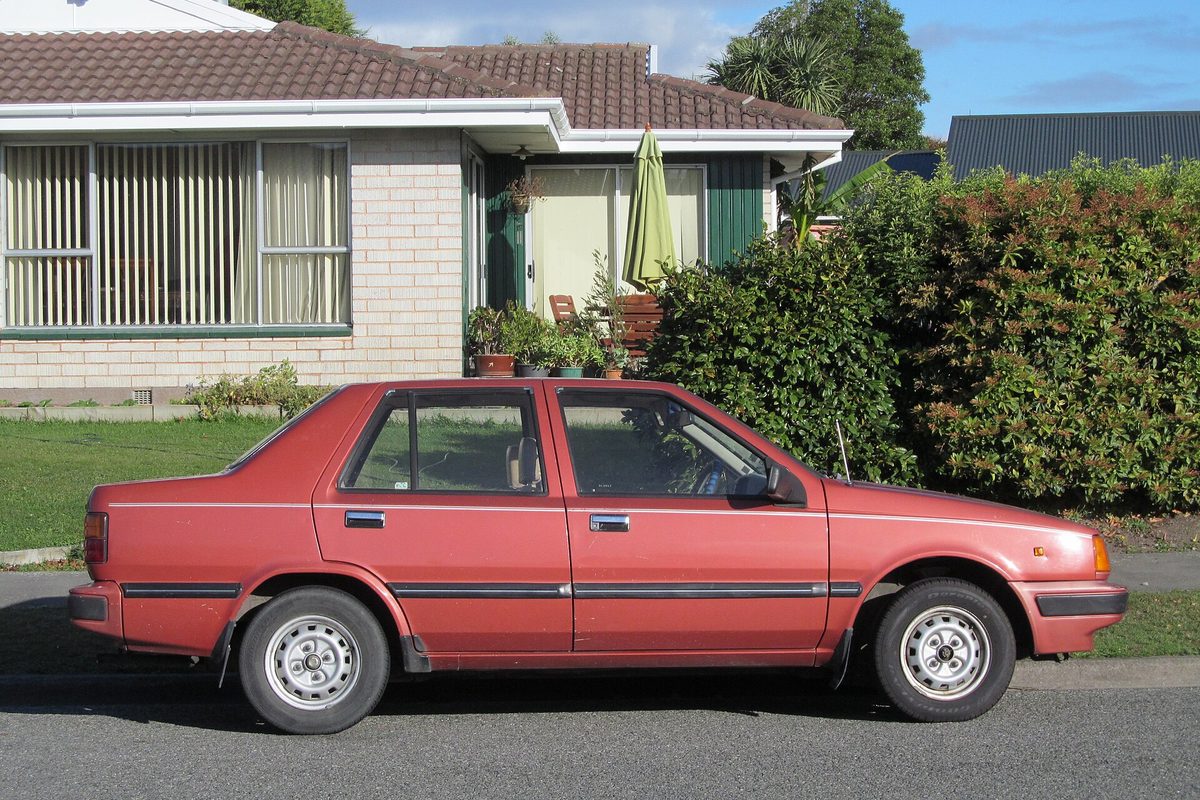 A faded red 1987 Hyundai Excel four-door sedan seen from the side, parked at a curb in front of a brick house and a tall hedge