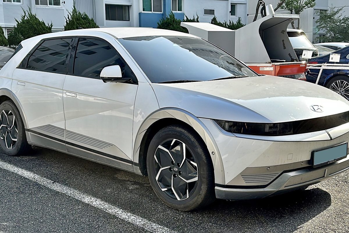 A white Hyundai Ioniq 5 crossover with dark lower trim and aero wheels, parked in a lot in front of apartment buildings