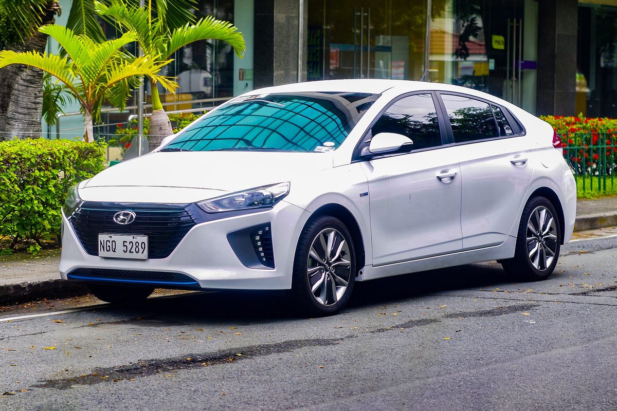 A white Hyundai Ioniq hybrid hatchback, front left view, parked on a city street beside palm trees and a glass-front building