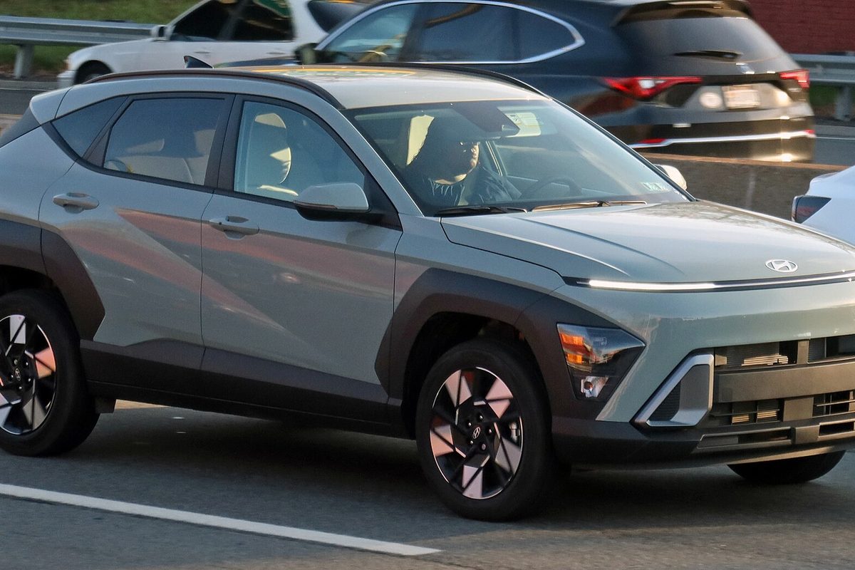 A pale green-gray 2024 Hyundai Kona with a full-width front light bar and two-tone wheels, driving on a highway