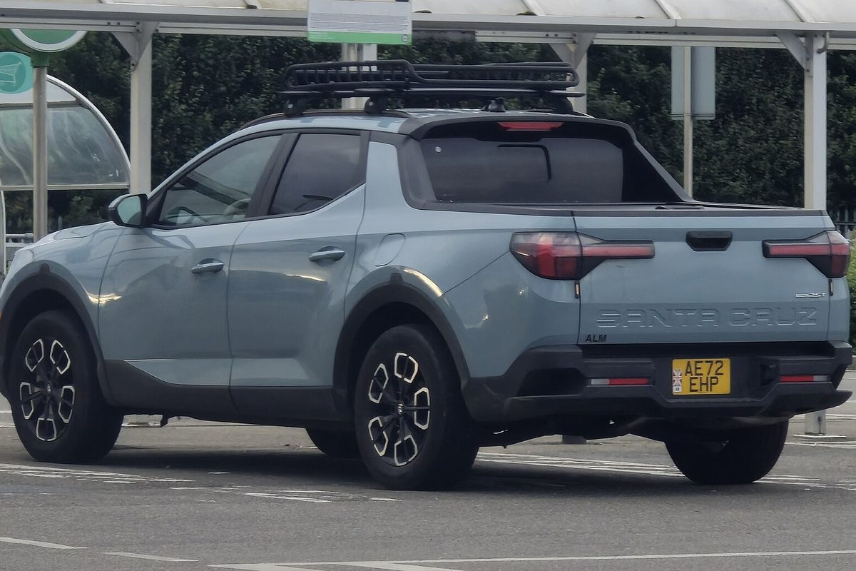 A pale gray-blue Hyundai Santa Cruz pickup seen from the rear left, with black alloy wheels and a roof rack, parked in a lot