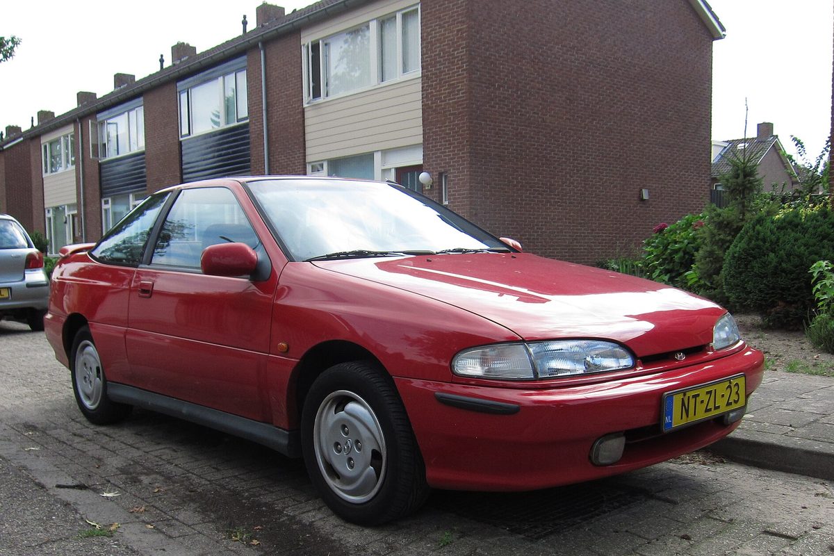 A red Hyundai Scoupe coupe with flush headlamps and silver wheel covers, parked on a brick street beside row houses
