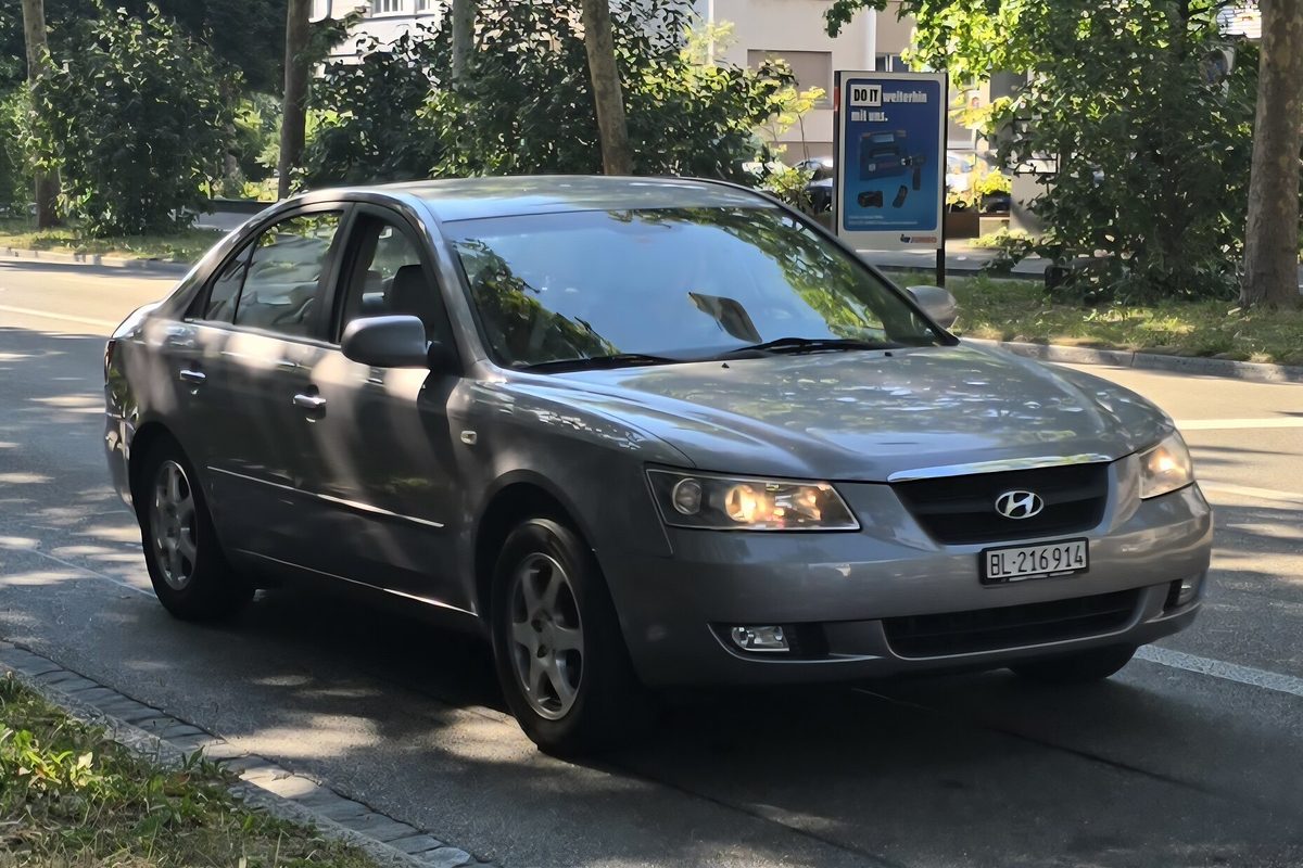 A gray Hyundai Sonata sedan with its headlights on, driving down a tree-lined city street