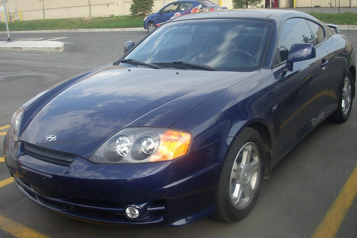A dark blue second-generation Hyundai Tiburon coupe with amber corner lights, parked in an empty lot
