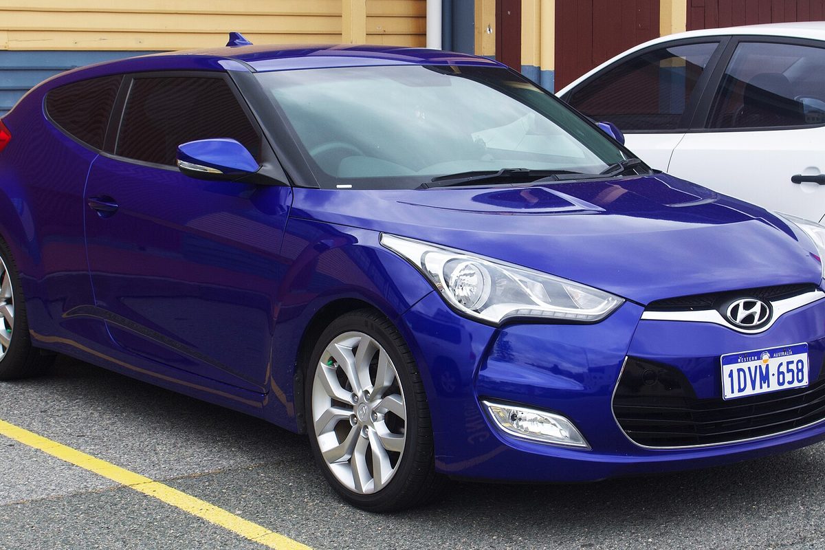A bright blue 2012 Hyundai Veloster hatchback parked in a lot with yellow lines, a white car beside it