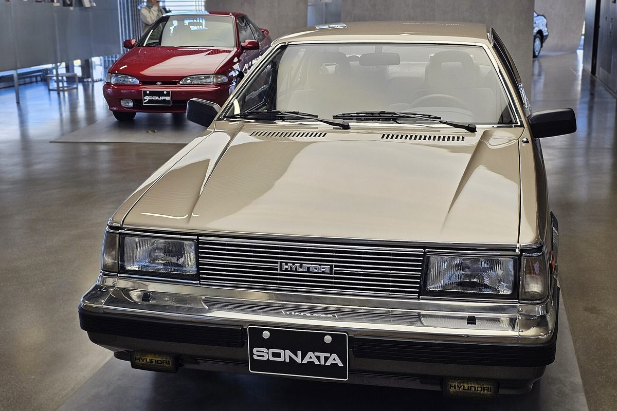 A gold first-generation (Y1) Hyundai Sonata 1.8 sedan on display at Hyundai Motorstudio Seoul, with a red Hyundai Scoupe parked behind it