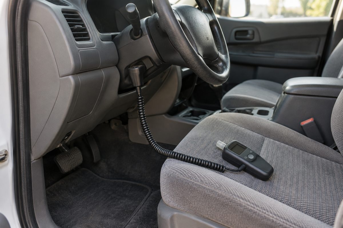 An ignition interlock handset on a coiled cord resting on a grey cloth driver's seat, its cable running up under the steering column