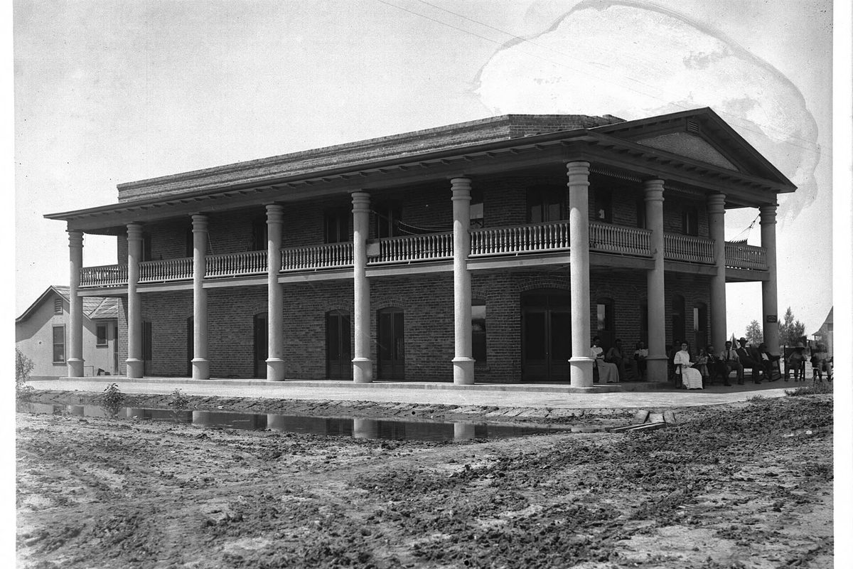 Black and white photograph from about 1910 of the two-story brick Hotel Imperial, with ten tall columns holding up a balcony, people on the porch and a muddy street in front