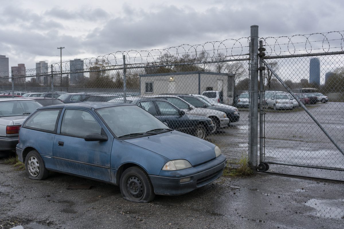 An old blue hatchback with a flat front tire parked inside a fenced tow lot, rows of cars and a small office behind it