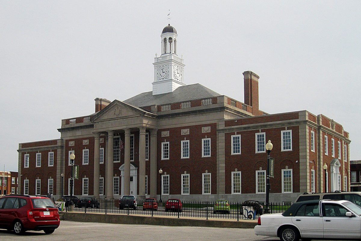 The red-brick Jackson County Courthouse in Independence, with a white clock tower, stone columns at the entrance and cars parked along the fence