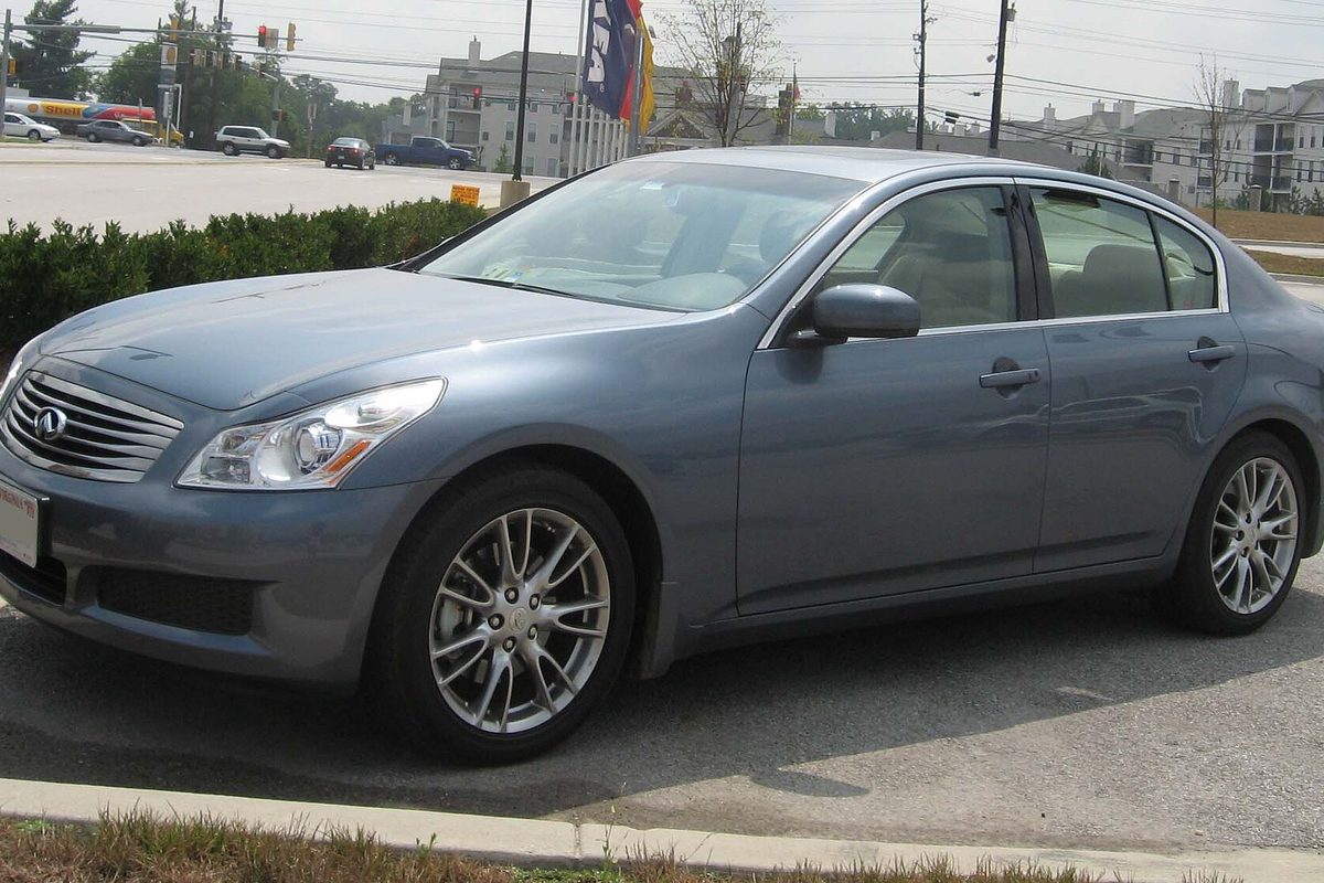 A blue-gray 2007 Infiniti G35 sedan parked beside a hedge at the edge of a parking lot