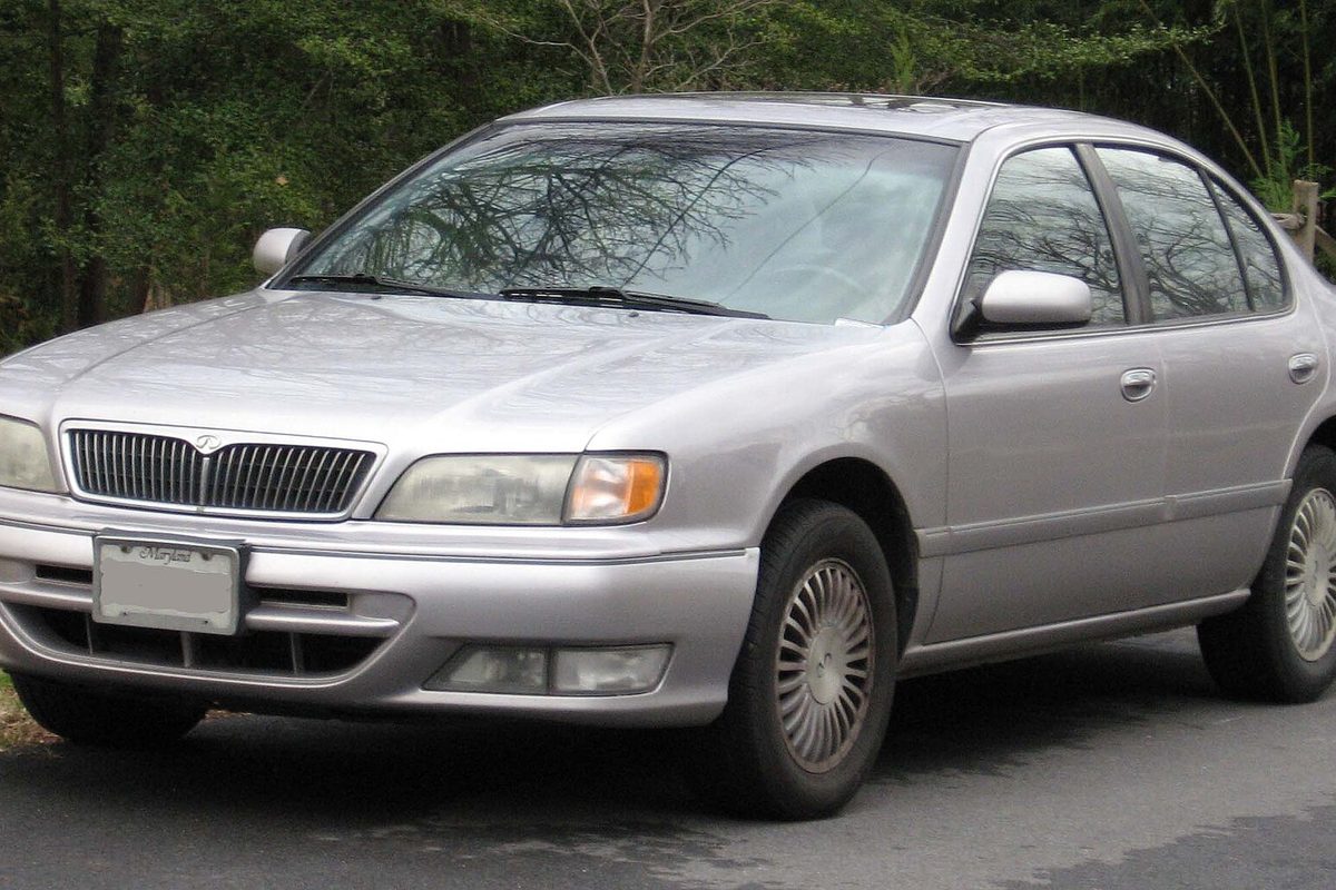 A silver-gray Infiniti I30 sedan with spoked wheel covers parked on a road in front of bare trees