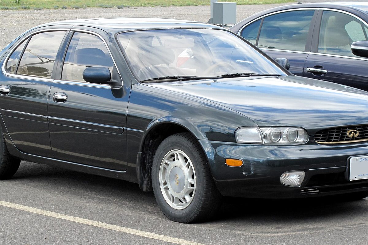 A dark green 1995 Infiniti J30 sedan with gold badges and white-and-silver wheels, parked in a lot beside another car