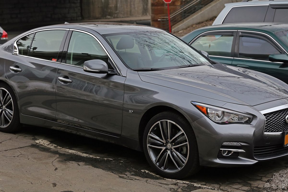 A gray 2014 Infiniti Q50 sedan parked on cracked pavement beside a green car