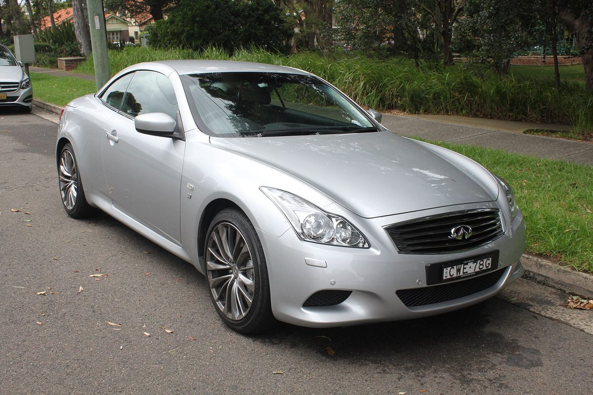A silver first-generation Infiniti Q60 convertible with its hard top raised, parked at a curb on a leafy street
