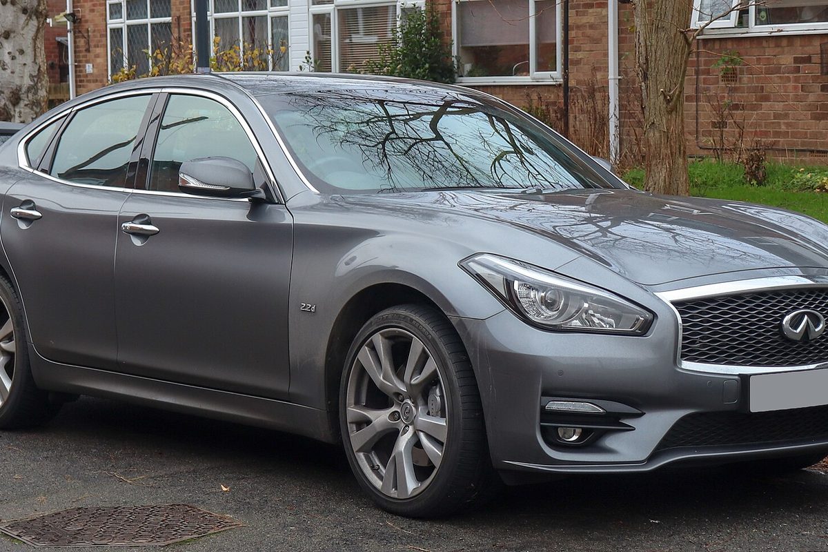 A dark gray Infiniti Q70 sedan with alloy wheels, parked on a wet street in front of a brick house