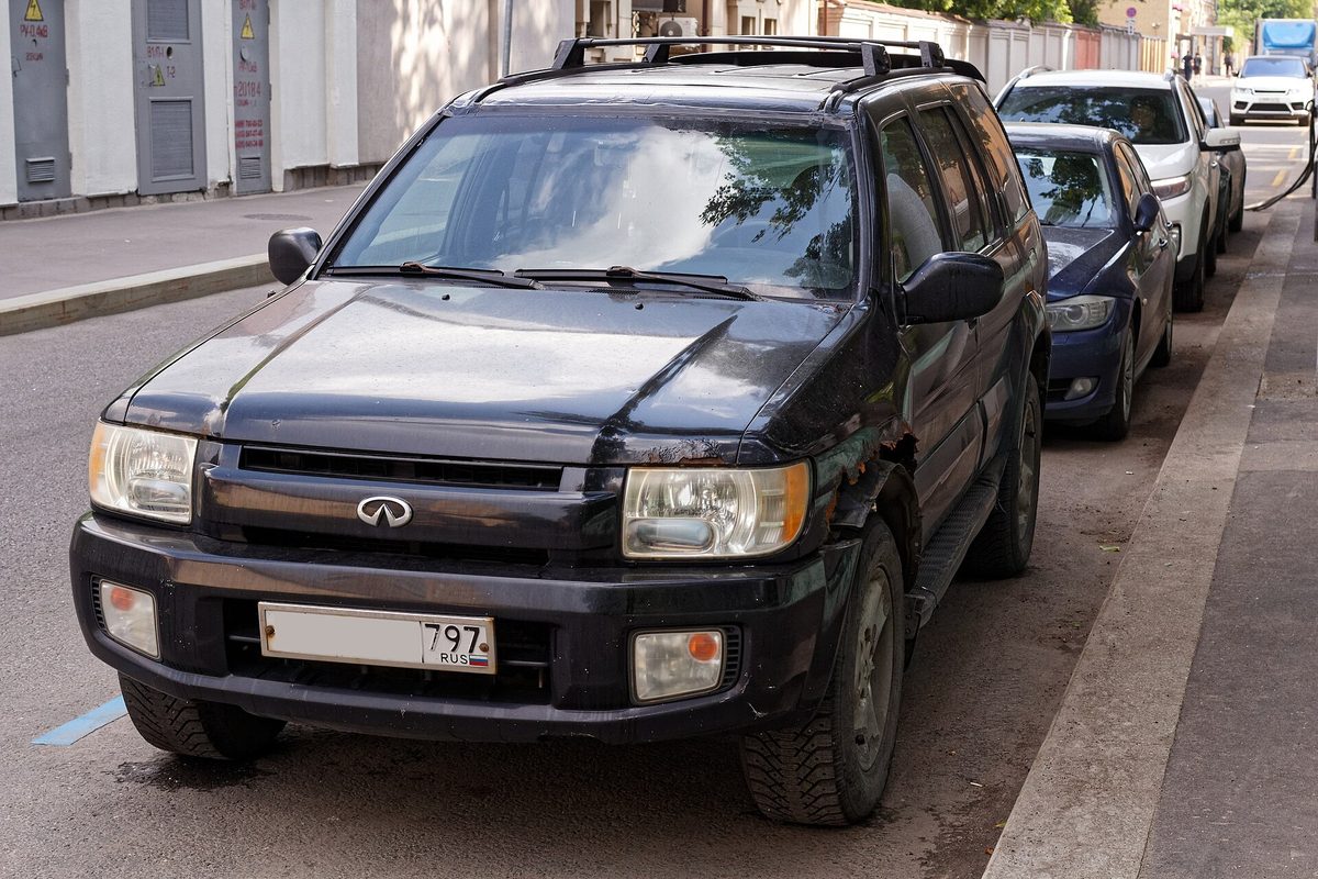 A black Infiniti QX4 SUV with roof rails and a scuffed front bumper parked at a city curb