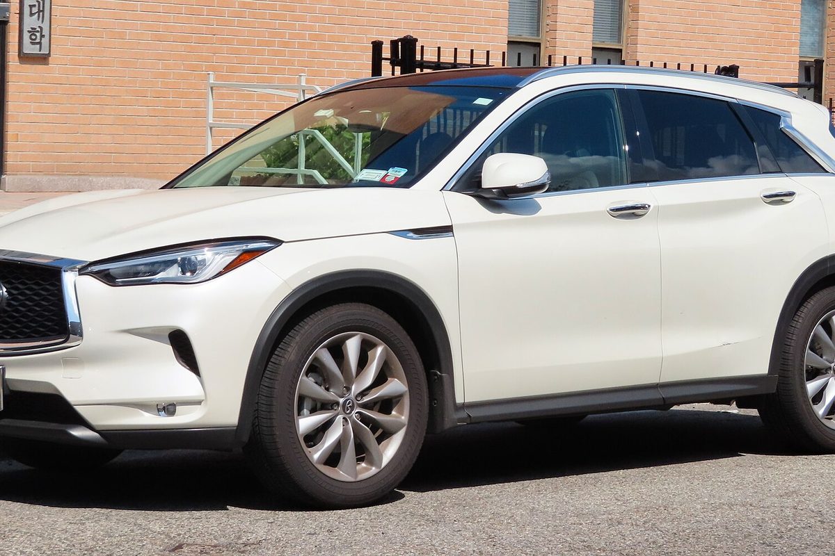 A pearl-white second-generation Infiniti QX50 crossover parked on a street beside a tan brick building