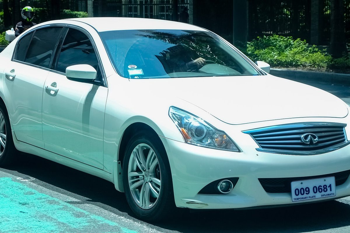 A white Infiniti G37x sedan, front three-quarter view, parked at a curb by a bike lane with a temporary diplomatic plate