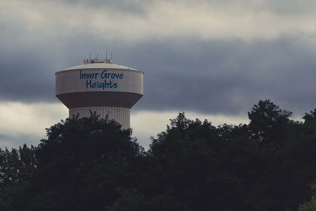 The Inver Grove Heights water tower, its name painted in blue script, rising above a dark line of trees under heavy gray clouds