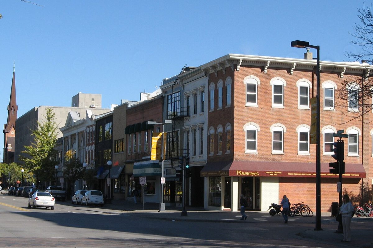 Brick storefronts on a sunny corner of Clinton Street in downtown Iowa City, a church steeple behind them and cars parked at the curb