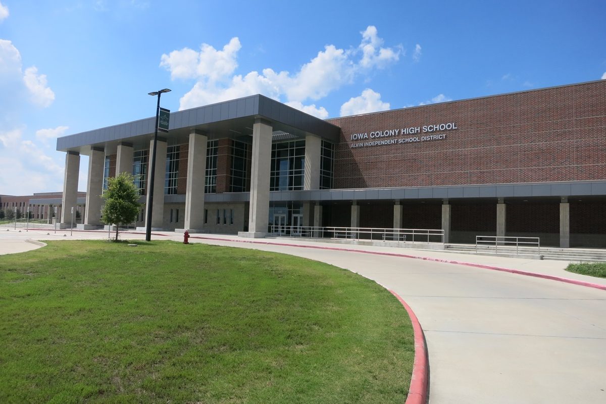 Iowa Colony High School, a red brick building with a tall columned entrance, behind a green lawn and a curving driveway under a blue sky