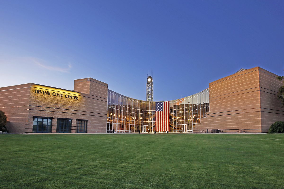 The Irvine Civic Center at dusk, with tan walls, a curved glass front behind a large American flag, a clock tower and a wide lawn