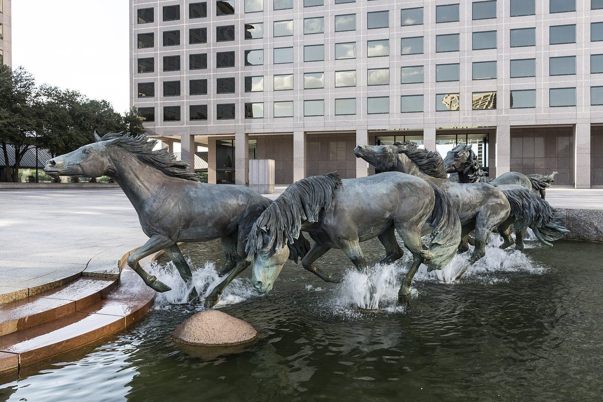 Bronze horses of the Mustangs of Las Colinas sculpture galloping through a fountain pool in front of a tall office building in Irving